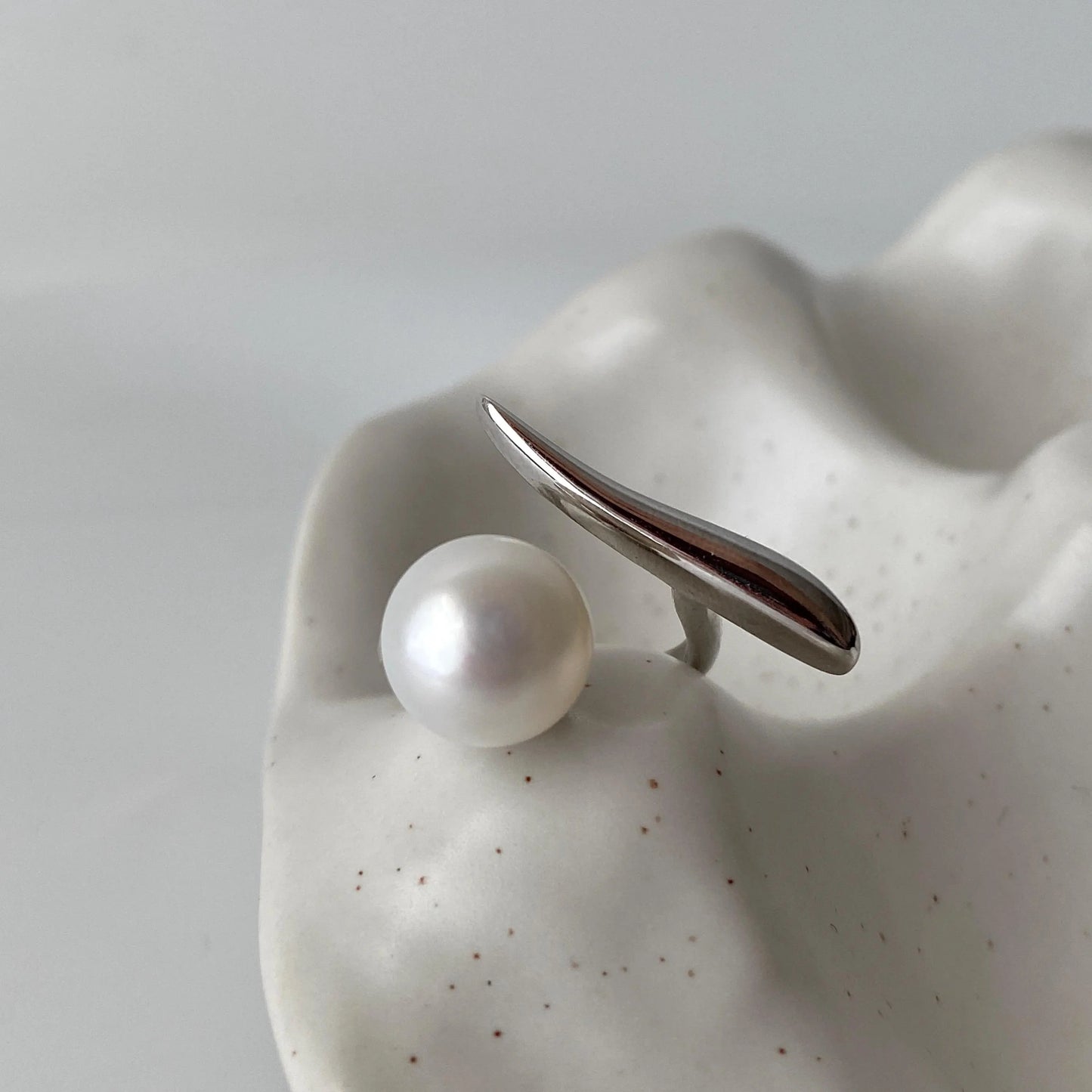 derMomentø - Handcrafted large pearl open ring with a naturally grown freshwater baroque pearl, set on a white rhodium-plated silver band. Display on a ceramic tray on a clean background.