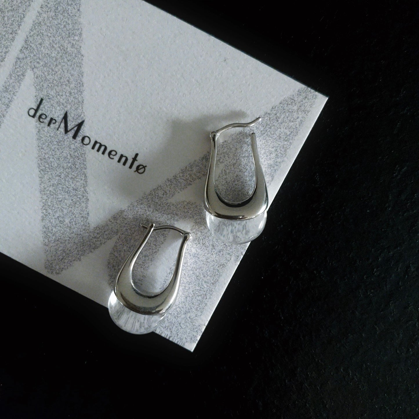 Close-up of bold silver hoop earrings with natural crystal quartz gemstones, displayed on brand card with name derMomentø, on a black background.