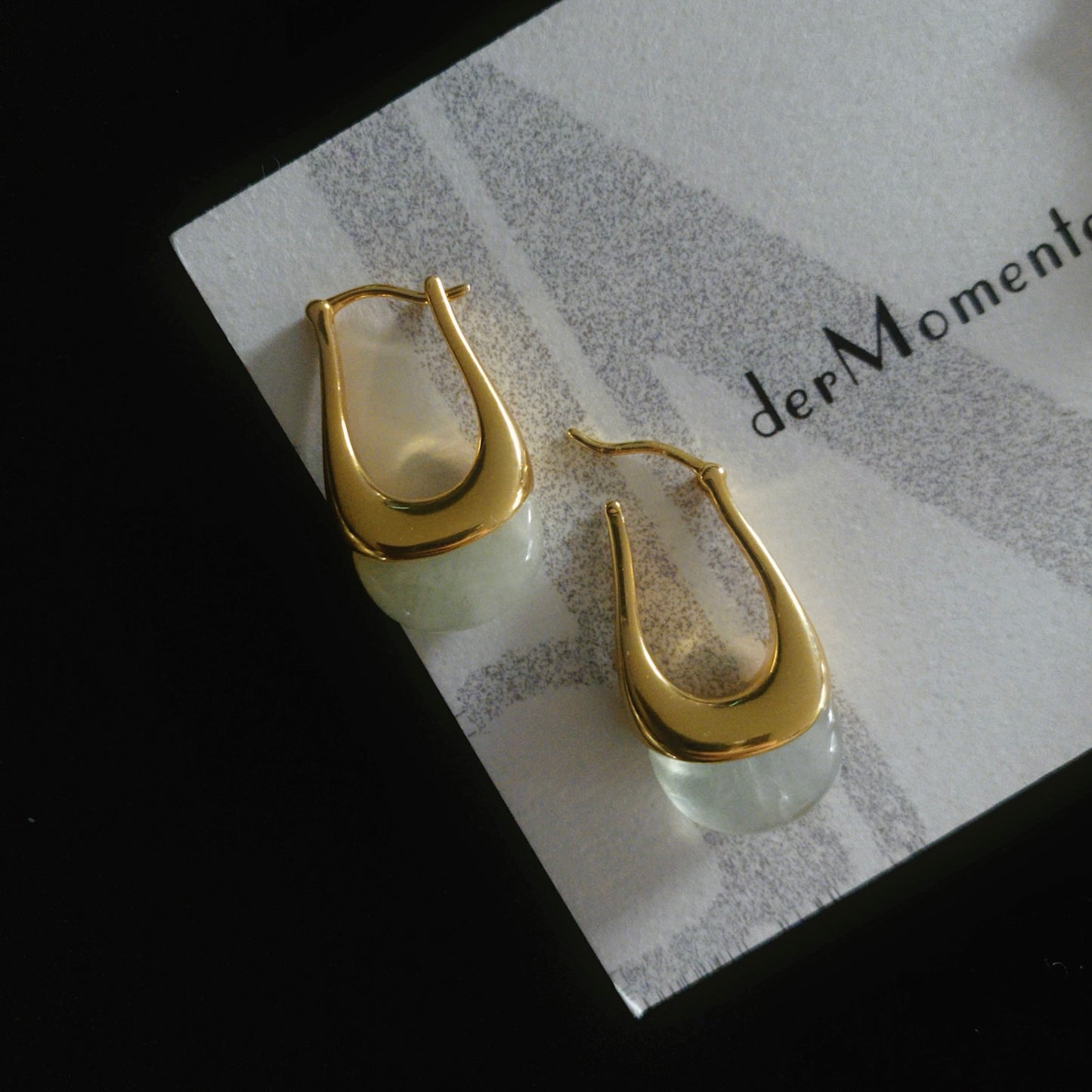 Close-up of chunky gold hoop earrings with natural green prehnite gemstones, displayed on brand card with name derMomentø, on a black background.
