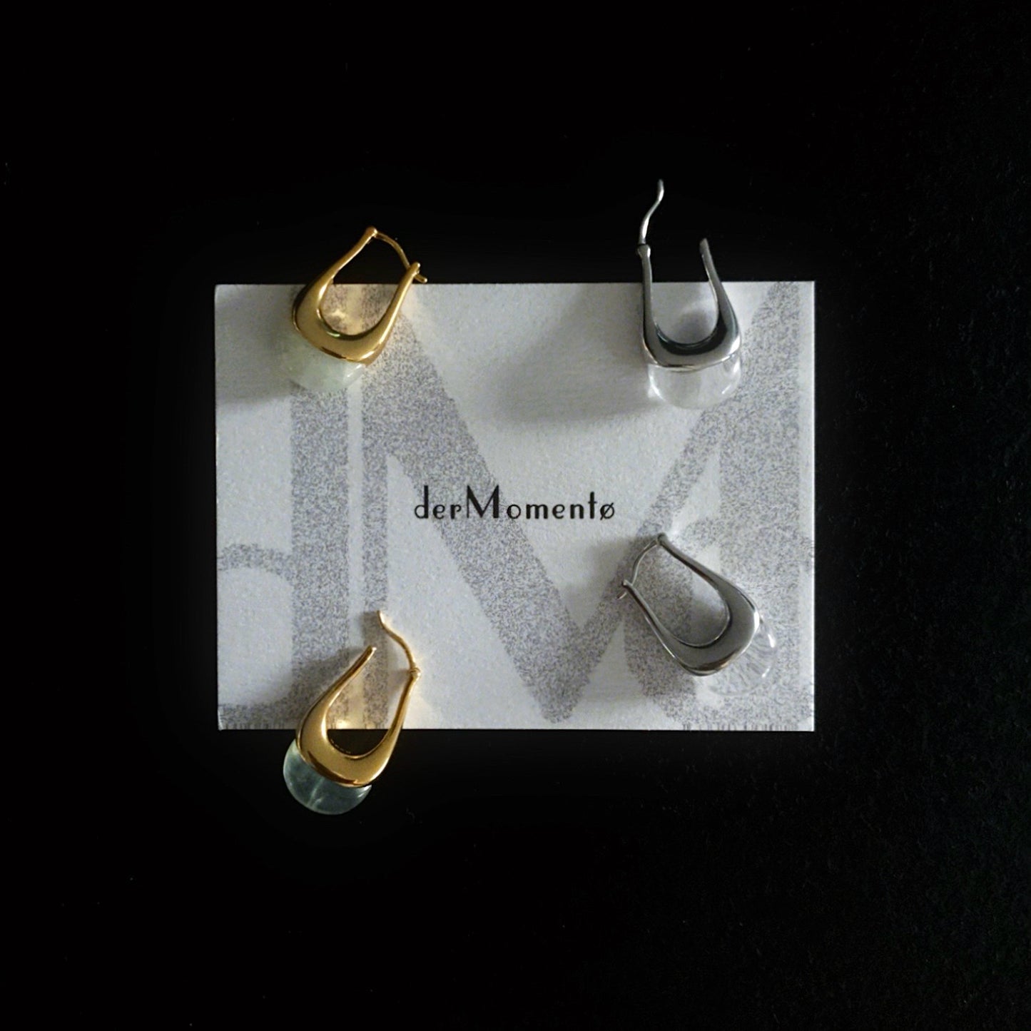 Two pairs of chunky silver and gold hoop earrings with natural green prehnite and clear quartz gemstones, modern minimalist jewelry design. Displayed on a brand card with name derMomentø, on a black background.