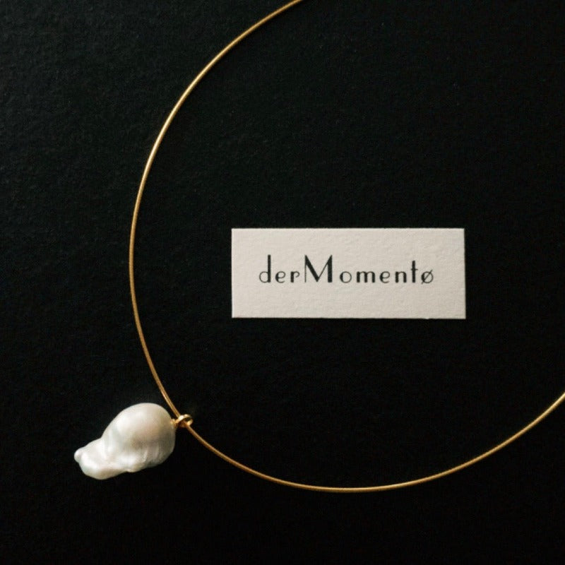 A close-up of Ariel Baroque Pearl Collar Necklace with an inverted natural baroque pearl pendant on an 18-karat gold vermeil silver collar choker, with brand name "derMomentø" at background.