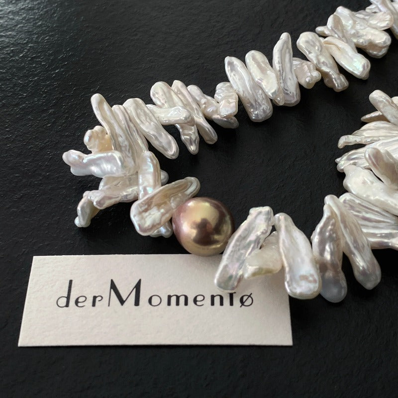 Close up of a cream biwa baroque pearl choker necklace with a uniquely coloured baroque pearl at its centre. Has brand "derMomentø" at background.