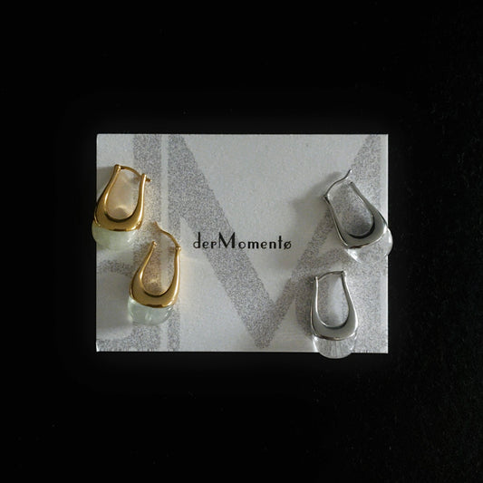 Chunky silver and gold hoop earrings with natural green prehnite and clear quartz gemstones, modern minimalist jewelry design. Displayed on a brand card with name derMomentø, on a black background.