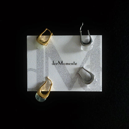 Two pairs of chunky silver and gold hoop earrings with natural green prehnite and clear quartz gemstones, modern minimalist jewelry design. Displayed on a brand card with name derMomentø, on a black background.