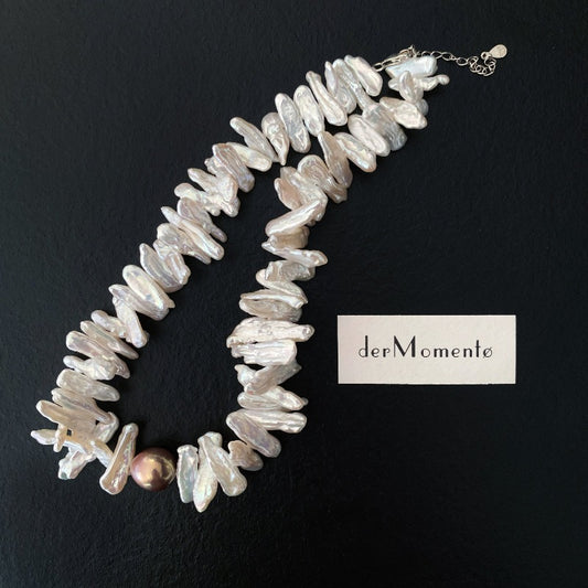 A choker necklace made from cream-toned biwa pearls, adorned with a uniquely coloured baroque pearl centrepiece and suspended by a sterling silver chain. Has brand "derMomentø" at background.