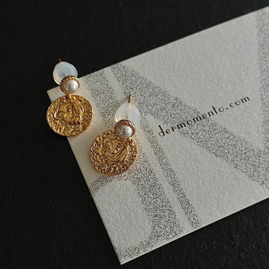 The Alwina Drop Stud Earrings - a mini pearl design (non-natural pearl) expertly enveloped in 18K gold vermeil silver and accented by tiny zircons and intricate tin relief detailing on each round tile. With dermomento.com at background.